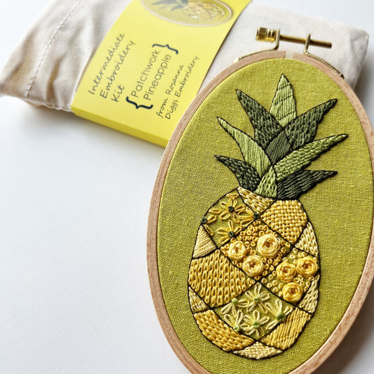 an embroidery of a pineapple stitched with many different embroidery stitches rests against an embroidery kit of the same design