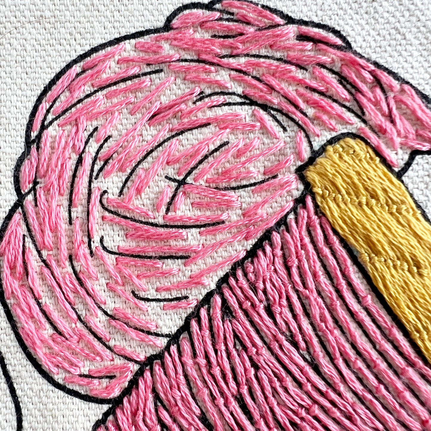 Close up of drop spindle and roving embroidered in pink on Rosanna Diggs Fiber Fluid tote. Roving has long straight stitches. Drop spindle has details in the woodwork design embroidered in a yellow gold.