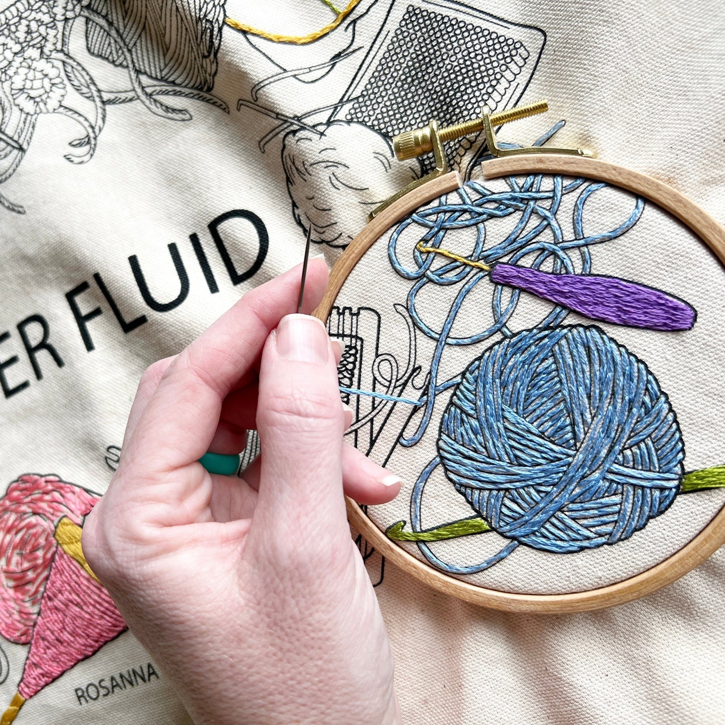 Close up of bag showing someone stitching a ball of yarn that is unraveled surrounded by crochet hooks. Yarn ball is embroidered in blue. Crochet hooks are green and purple. Four inch hoop surrounds area being worked on.