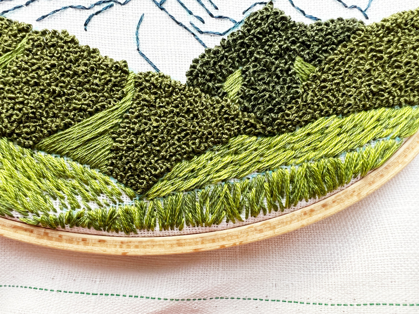 Up close photo of Hurricane Ridge Embroidery. Stitch details in green highlight trees, hills and grass. Mountain can be seen in the background.