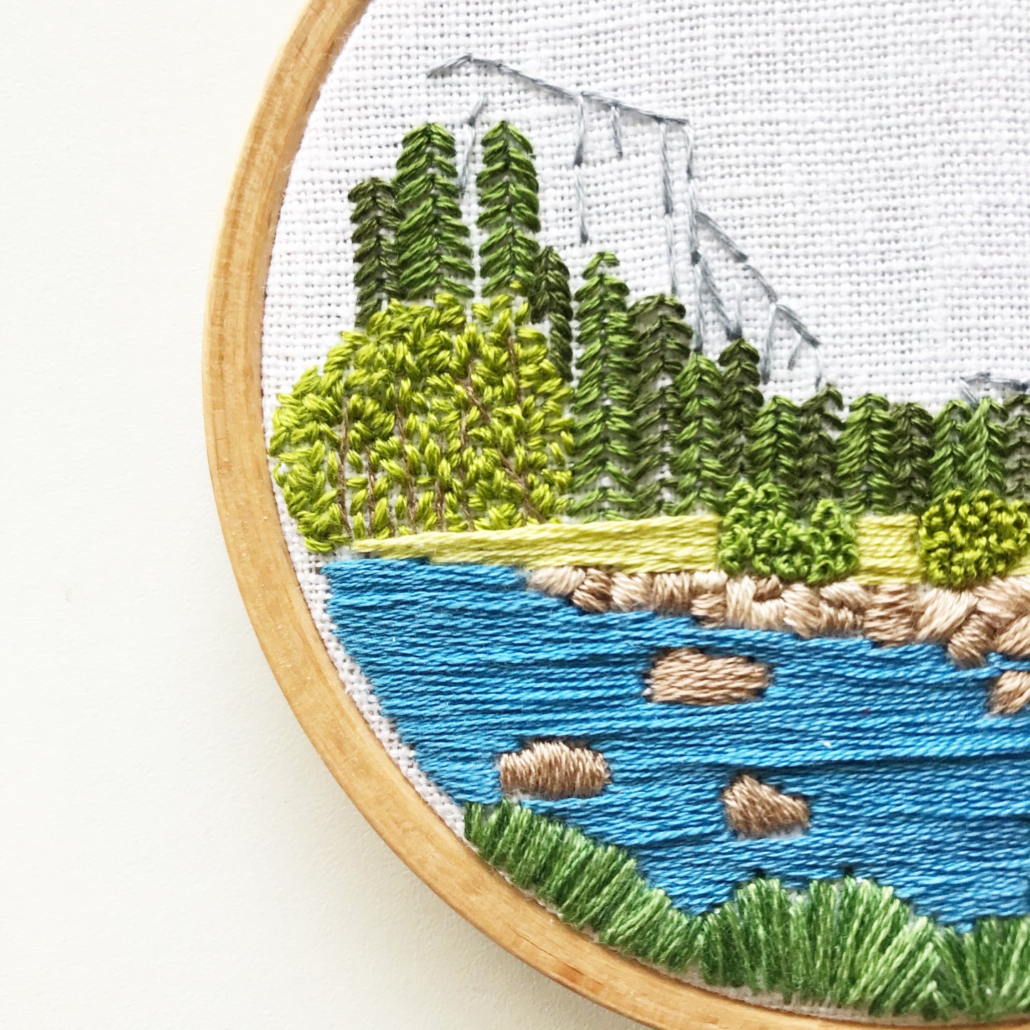 Close up of Yosemite Valley Embroidery Kit.  Stitch detail of blue water surrounded by trees and shrubs in shades of green.  Mountains of the valley are in the background.