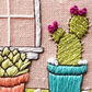 Close up of cactus showing spines and flowers in a teal pot with white framed window on peach linen.  Cactus detail of Cozy Cacti by Rosanna Diggs Embroidery.