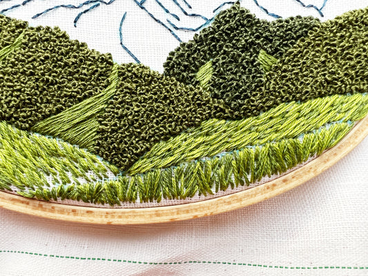 Up close photo of Hurricane Ridge Embroidery.  Stitch details in green highlight trees, hills and grass.  Mountain can be seen in the background.