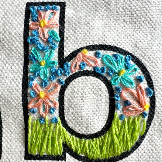 Close up of stitch detail on Love Your Neighbor tote.  Letter has long grass stitched at the base and the rest is filled with flowers.